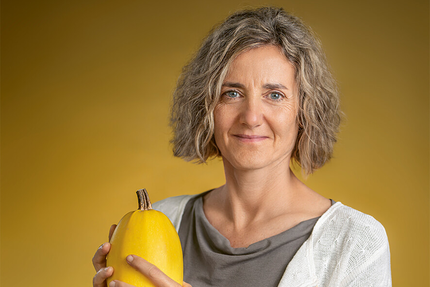 Die Ernährungsberaterin Sibylle Birnstiel schaut in die Kamera und hebt einen Kürbis als Zeichen der saisonalen Ernährung hoch.