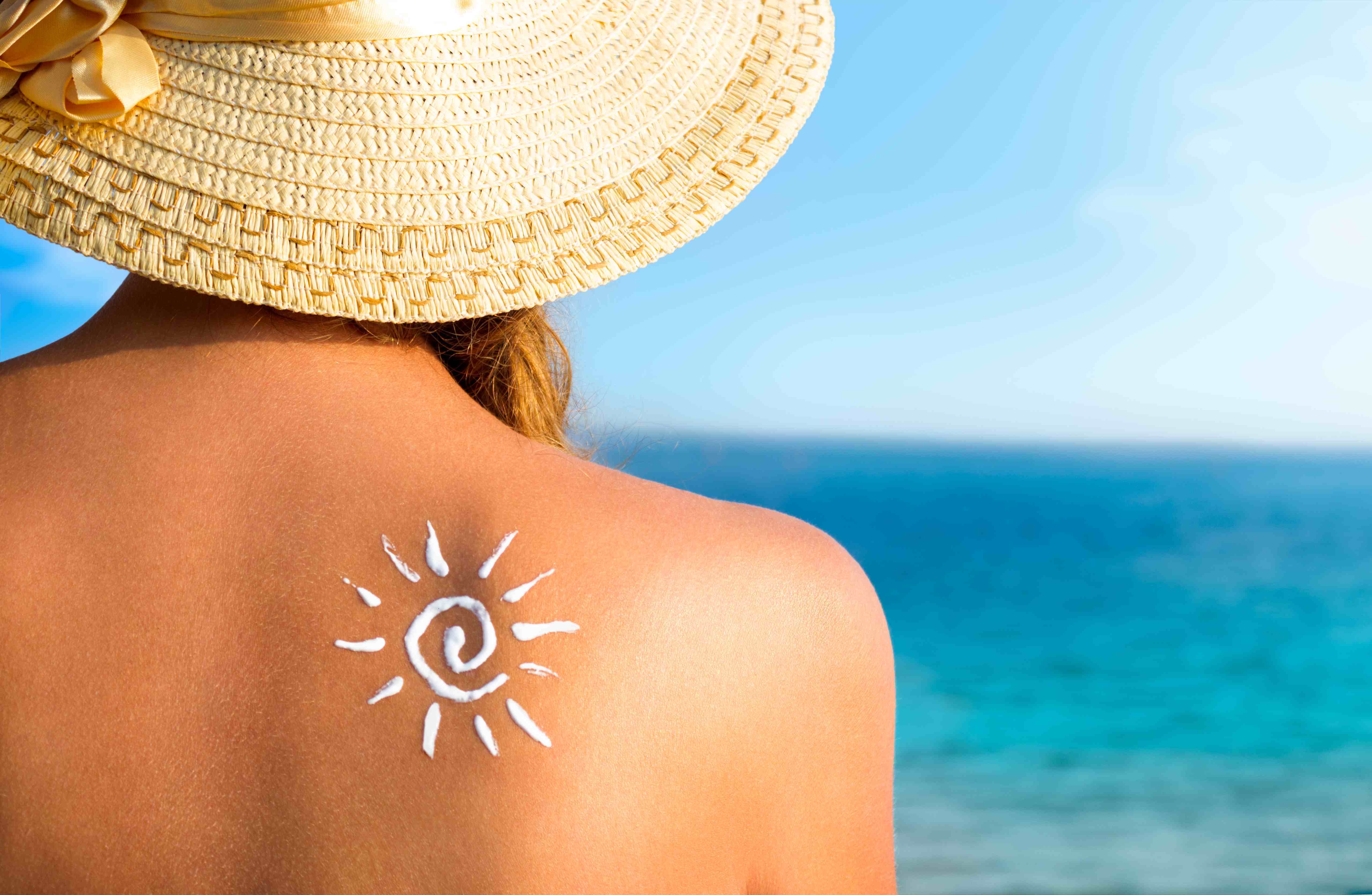 Frau mit Sonnenhut und Sonnencreme auf dem Rücken am Strand aufs Meer blickend